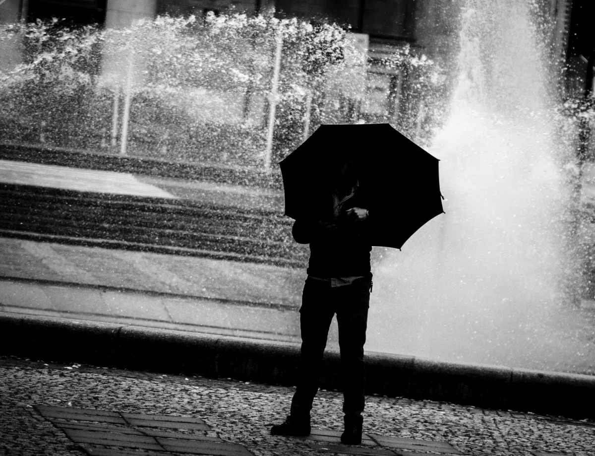 Figure holding a black umbrella before a fountain spray